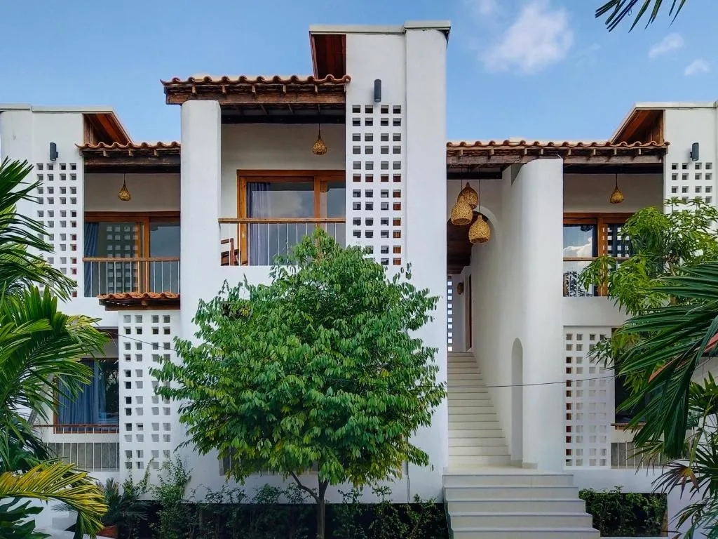 Two-storey white building at Canary Guesthouse with terracotta roof tiles, breeze-block panels, exterior stairs and a front garden tree