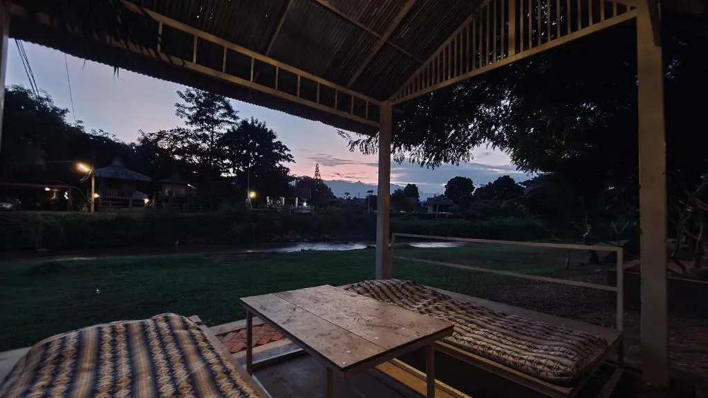 Covered riverside sala at Canary Guesthouse at dusk with floor cushions, a low table and lights across the water