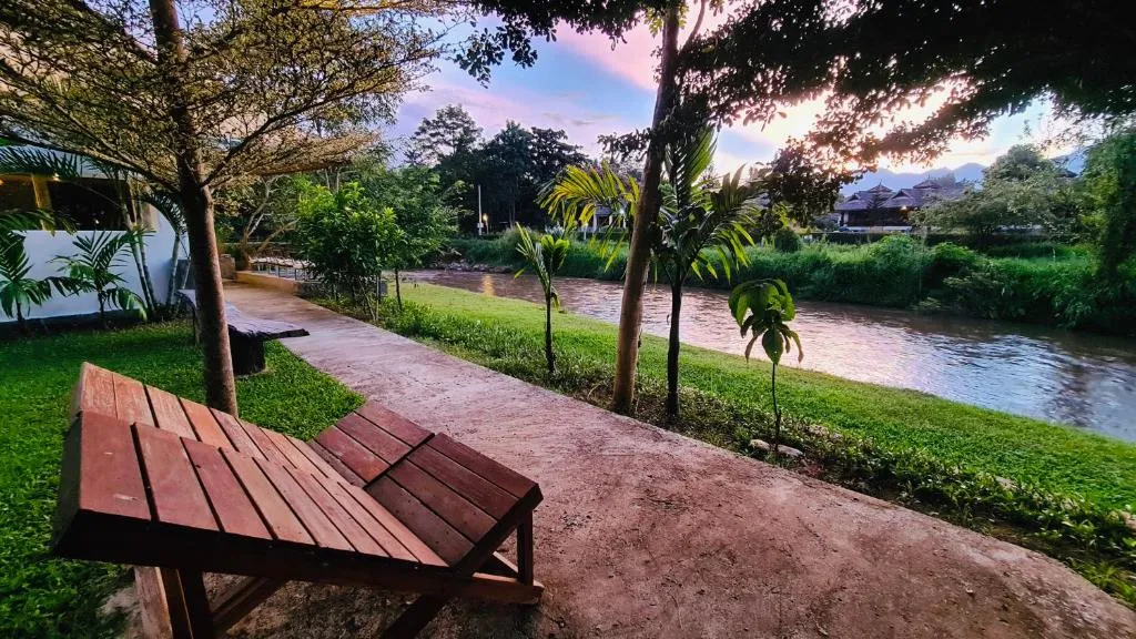 Red-dirt riverside path with a wooden bench and lawn running along the Pai River at Canary Guesthouse at dusk