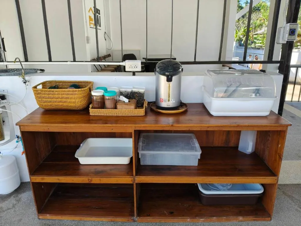 Self-service coffee and tea station at Canary Guesthouse with a hot-water urn, mugs and supplies on a wooden shelf