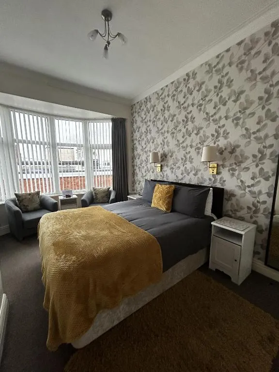 Bay-window room at The Inglewood Hotel with a large bed, mustard throw, grey-leaf feature wallpaper and a small sofa by the window